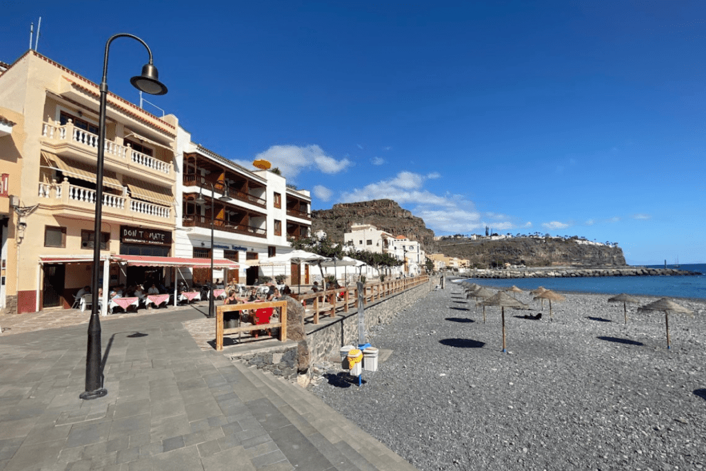 Restaurants an der Promenade von Playa De Santiago La Gomera zeigt auch einen felsigen Strand mit Sonnenschirmen und blauem Meer, einer der besten Strände von La Gomera 