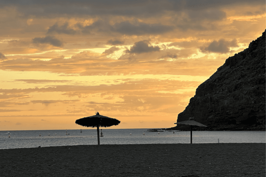 Sonnenuntergang an der Playa de San Sebastian mit der Silhouette eines Sonnenschirms und schwarzen Klippen rechts, einer der besten Strände in San Sebastian De La Gomera, Kanarische Inseln, Spanien
